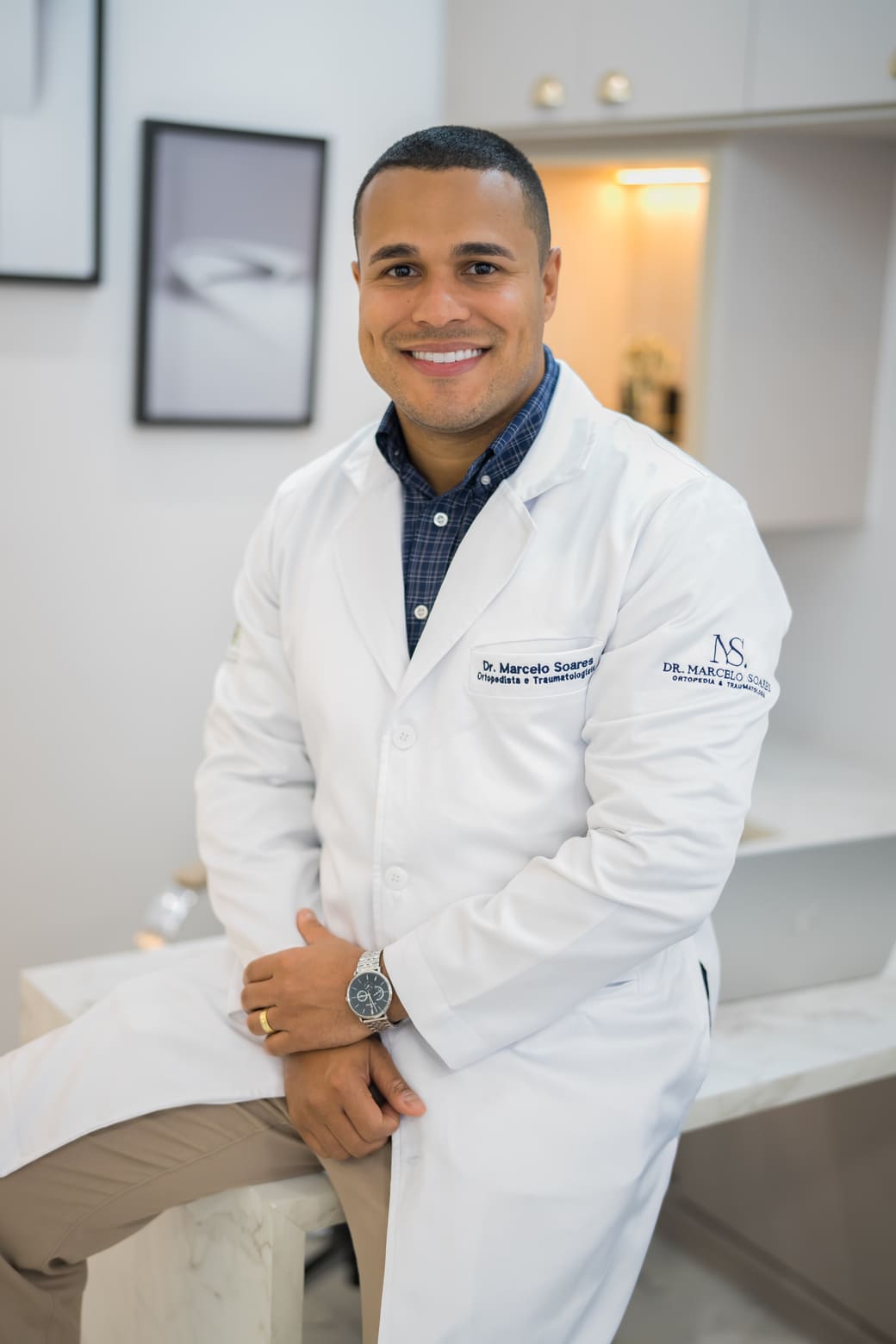 Dr. Marcelo Soares, ortopedista, sorrindo de jaleco branco bordado apoiado sobre bancada de mármore em consultório moderno, quadro abstrato ao fundo, iluminação suave, fotografia profissional ultra-realista