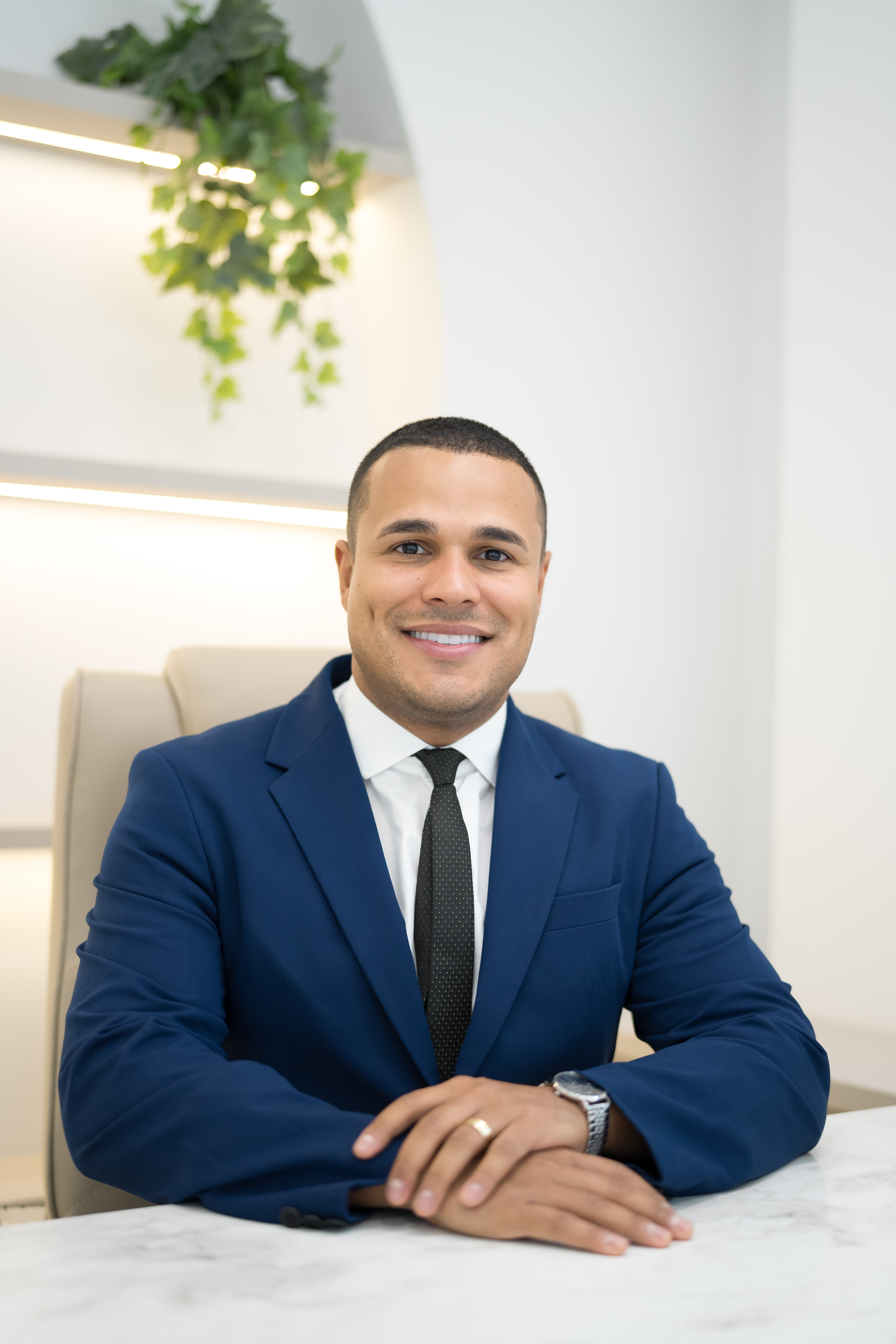 Dr. Marcelo Soares, ortopedista, sorrindo de terno azul-marinho, mãos entrelaçadas sobre mesa de mármore branco, fotografia profissional ultra-realista